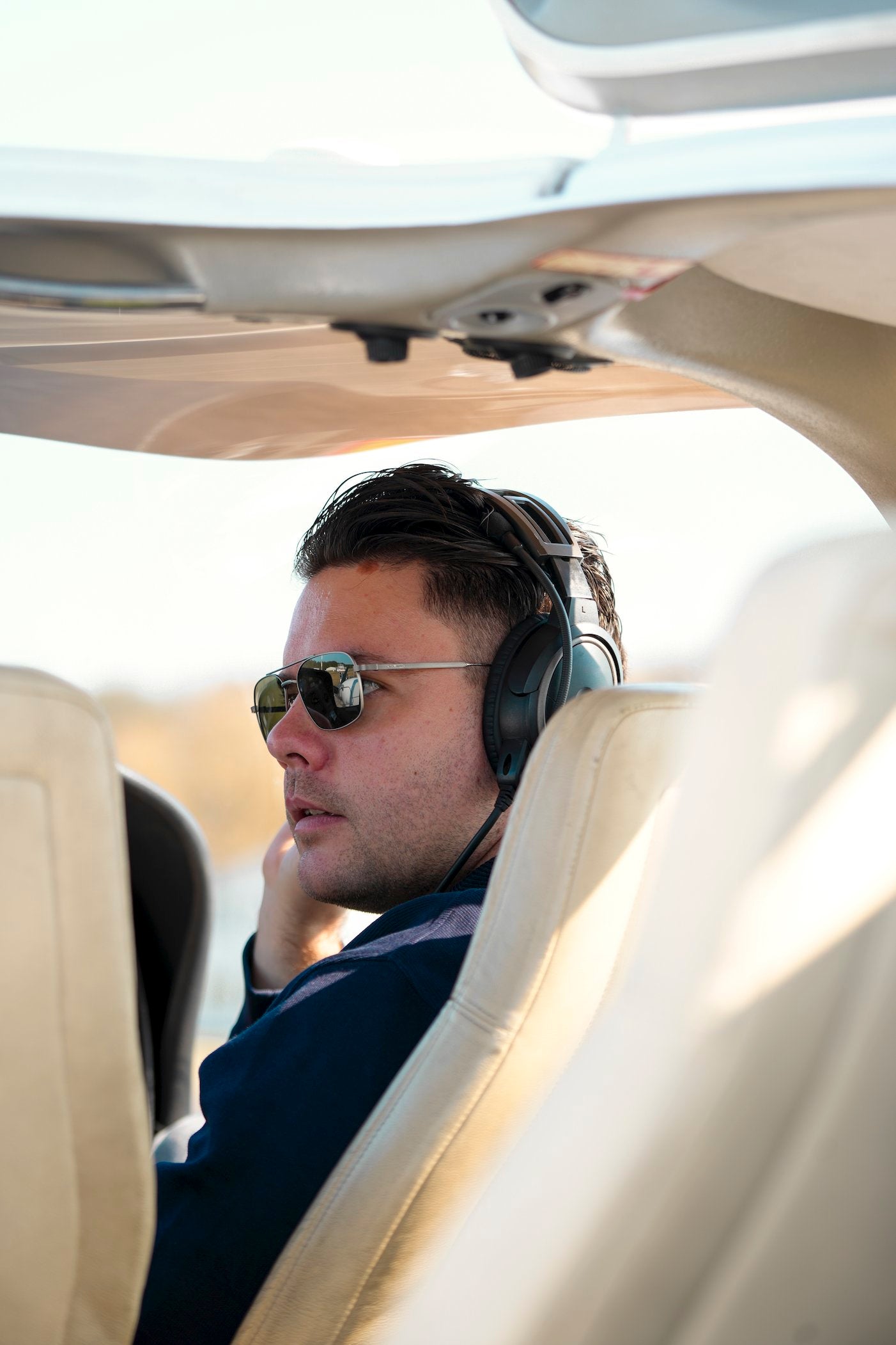 Man wearing aviator sunglasses and a headset seated in an aircraft cockpit, looking to the side.