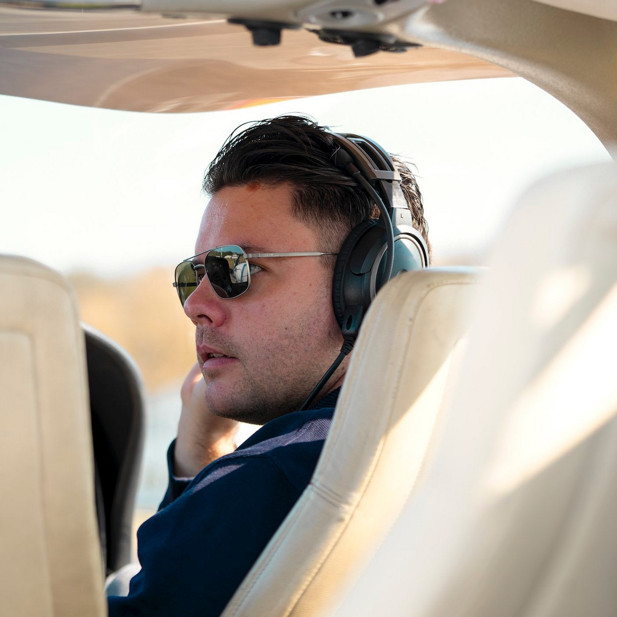 Lifestyle photo of a man wearing silver aviator-style sunglasses and a pilot headset while seated in an aircraft cockpit.
