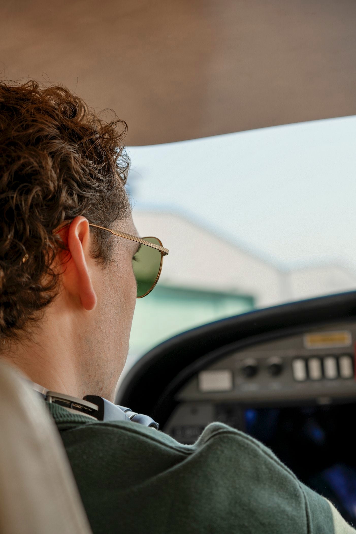 Close-up profile of a man wearing gold aviator sunglasses with green lenses, seated inside an aircraft cockpit.