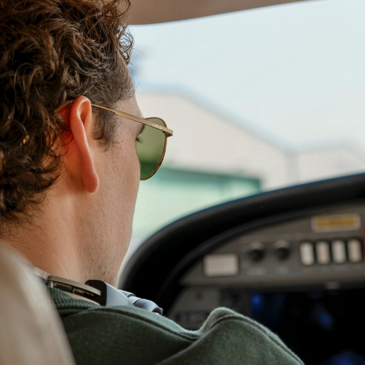 Man wearing gold aviator sunglasses with green-tinted lenses, seen from behind in profile, seated in a small aircraft cockpit looking out toward a hangar.