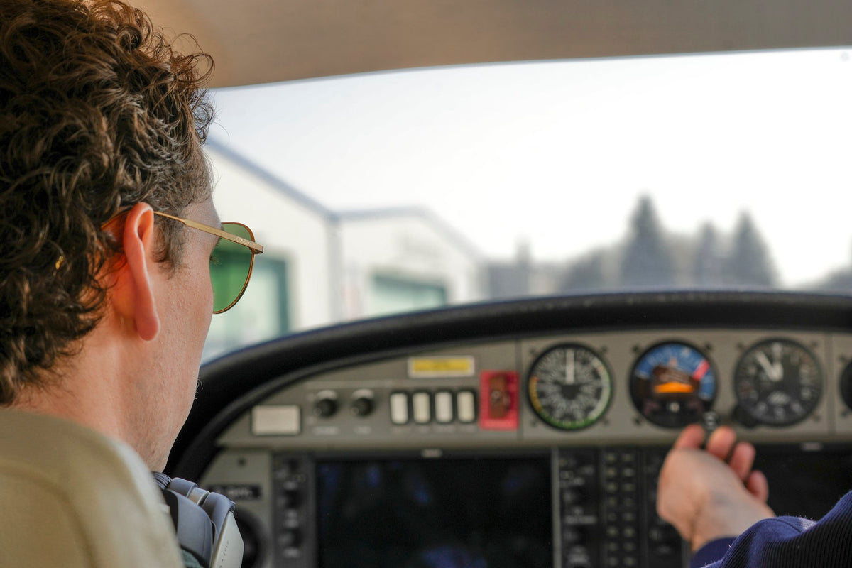 Close-up of a man wearing gold aviator sunglasses with green lenses, seen from behind inside an aircraft cockpit with instrument panel visible.