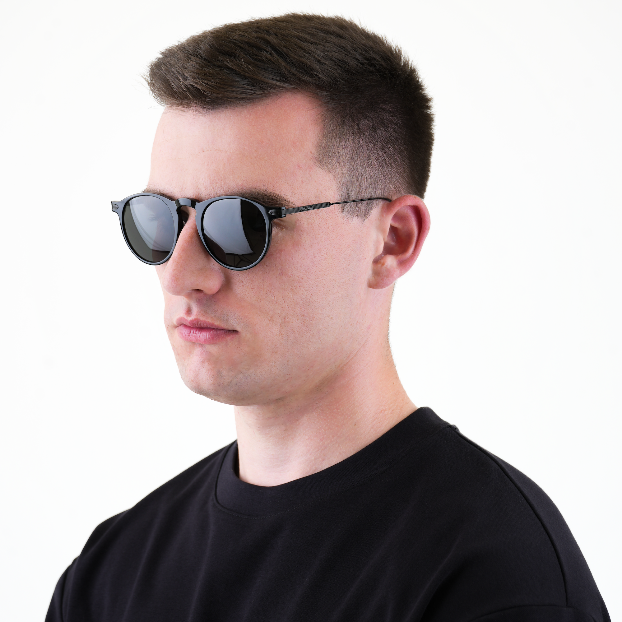 A man wearing black round sunglasses with dark lenses and thin metal frames, dressed in a black crewneck shirt against a plain white background.
