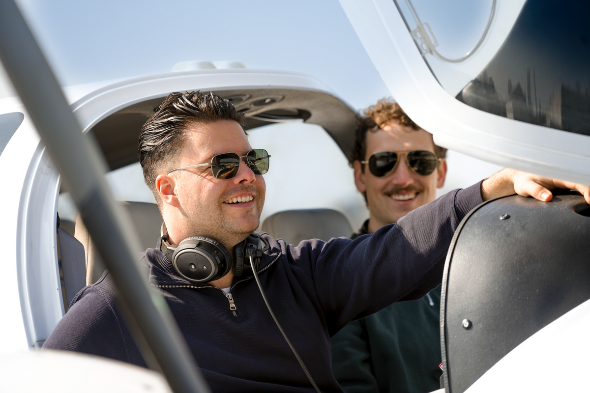 Two men wearing aviator sunglasses and a headset smiling inside the cockpit of a small aircraft, one with his arm extended.